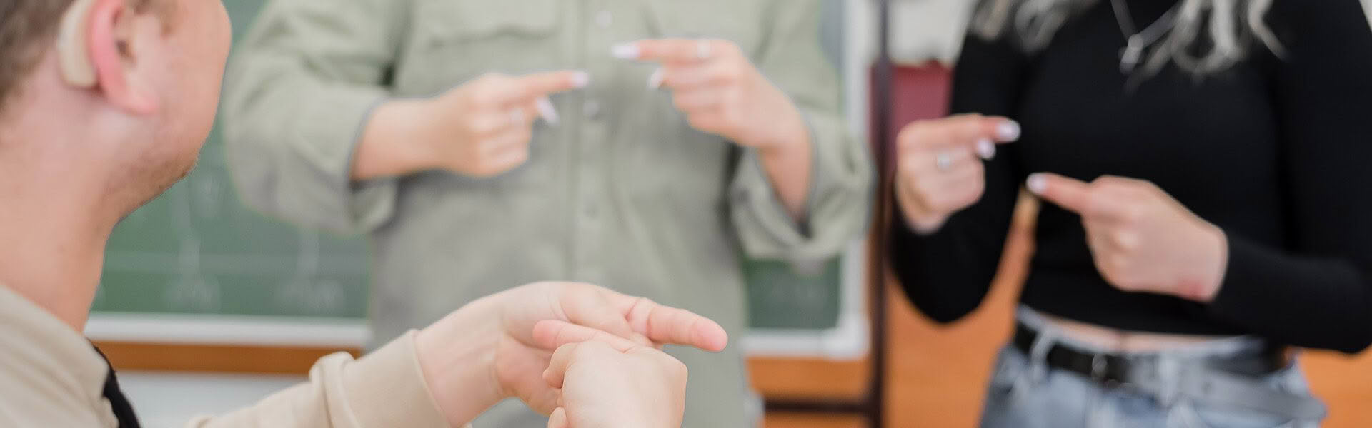 A man wearing a hearing aid is teaching the sign ABOUT in NZSL to 2 woman, they are in a classroom
