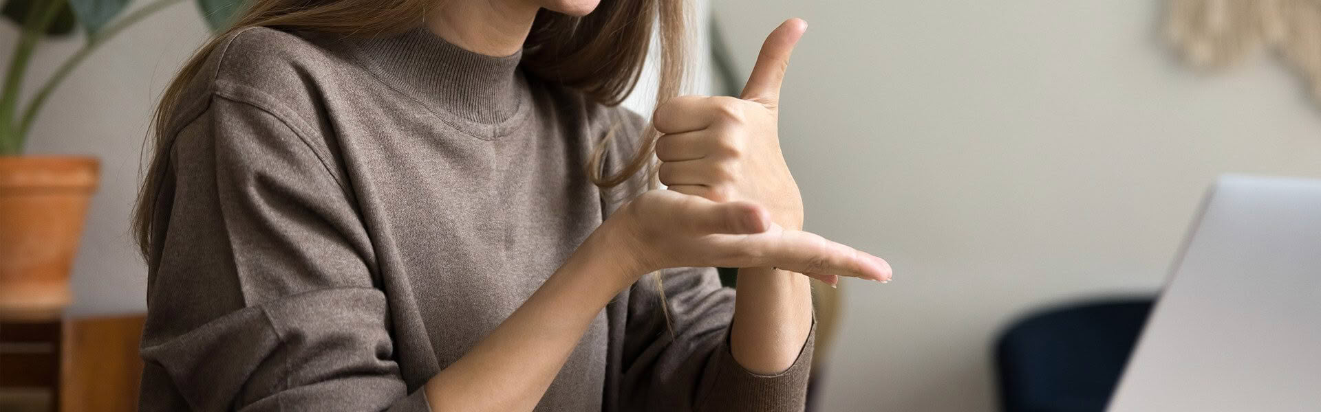 A woman wearing a light brown top is sitting in front of a laptop computer, she is signing KIA ORA in NZSL to someone on the computer screen